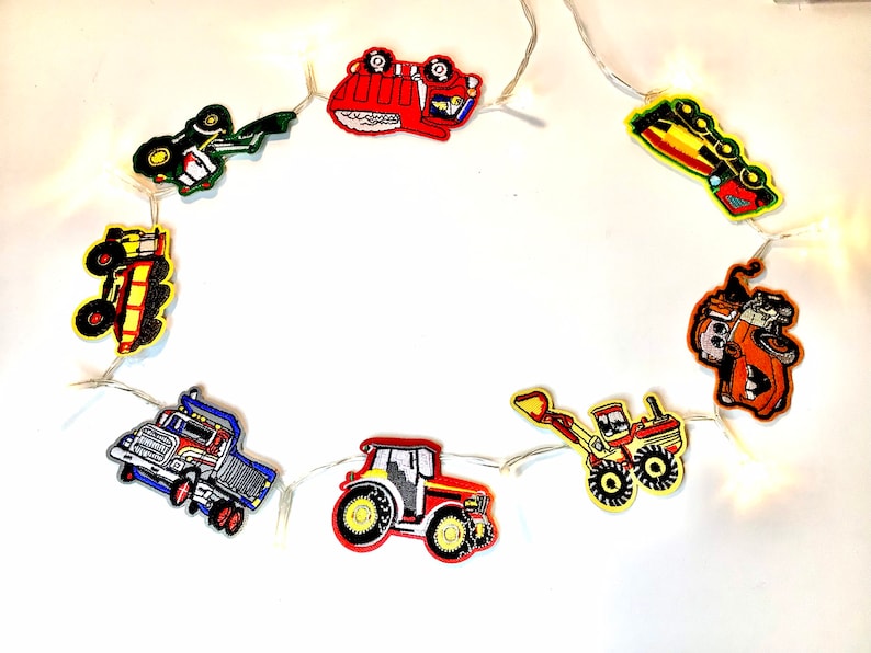 May include: A string of white fairy lights with colourful felt construction vehicle cut-outs. The vehicles include a dumper truck, a tractor, a bulldozer, a cement mixer, and a digger.