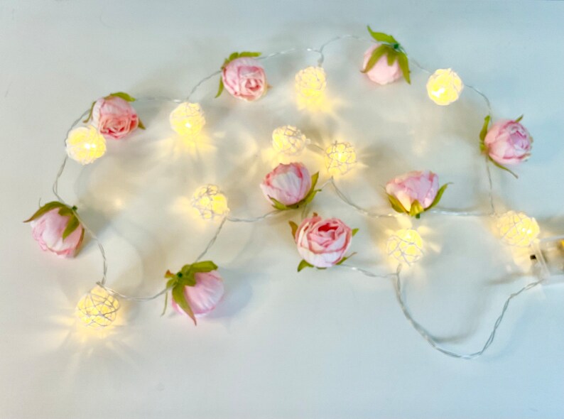 May include: A string of fairy lights with pink flower accents. The lights are white and the flowers are made of fabric.