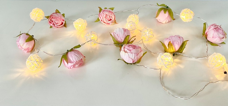 May include: A string of fairy lights with pink fabric flowers and white wire cage bulbs. The lights are strung together with a silver wire.