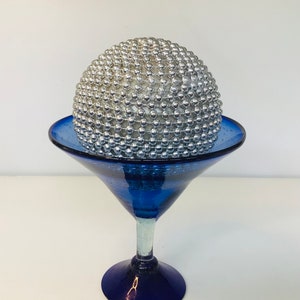 K&ouml;nnte beinhalten: Eine silberne Kugel, die mit kleinen Perlen bedeckt ist, sitzt auf einem blauen Glas-Martini-Glas.