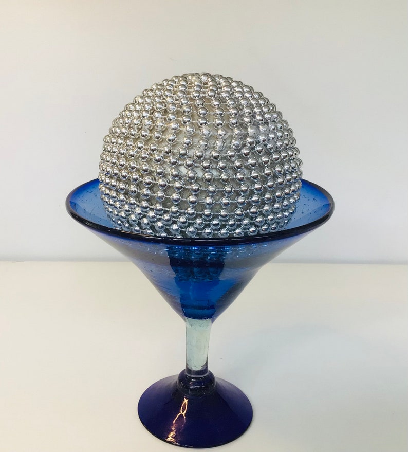 K&ouml;nnte beinhalten: Eine silberne Kugel, die mit kleinen Perlen bedeckt ist, sitzt auf einem blauen Glas-Martini-Glas.
