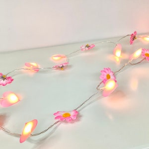 Può includere: Una ghirlanda di luci con lampadine a forma di fiori rosa. Le luci sono infilate su un filo argentato e sono illuminate da una luce bianca calda.
