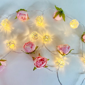May include: A string of white fairy lights with pink fabric flowers and white woven ball lights. The lights are strung together with a clear wire.