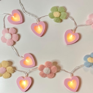 Op de afbeelding: Een slinger van decoratieve lampjes met roze hartvormige lampjes en vilten bloemen in verschillende kleuren. De bloemen zijn wit, roze, geel, blauw en paars. De lampjes zitten aan een witte draad.