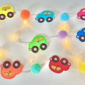 Puede incluir: Una colorida guirnalda con temática de coches, con recortes de fieltro de coches en varios colores, como amarillo, azul y rosa. La guirnalda también cuenta con pompones y pequeñas luces LED blancas cálidas, creando una decoración festiva.