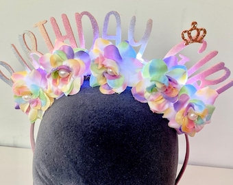 Puede incluir: Una diadema festiva de cumpleaños con flores de tela de colores pastel con centros de perlas. La diadema tiene la palabra "birthday" en letras brillantes y una pequeña corona dorada. Las flores están dispuestas en la parte superior.