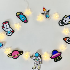 May include: A string of white fairy lights with various space-themed felt patches, including a rocket, astronaut, UFO, and planet. The lights are strung together with small white stars.