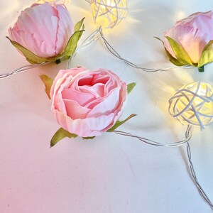 May include: Pink fabric rose flower buds with white woven ball lights on a white background. The lights are strung together with a silver wire.