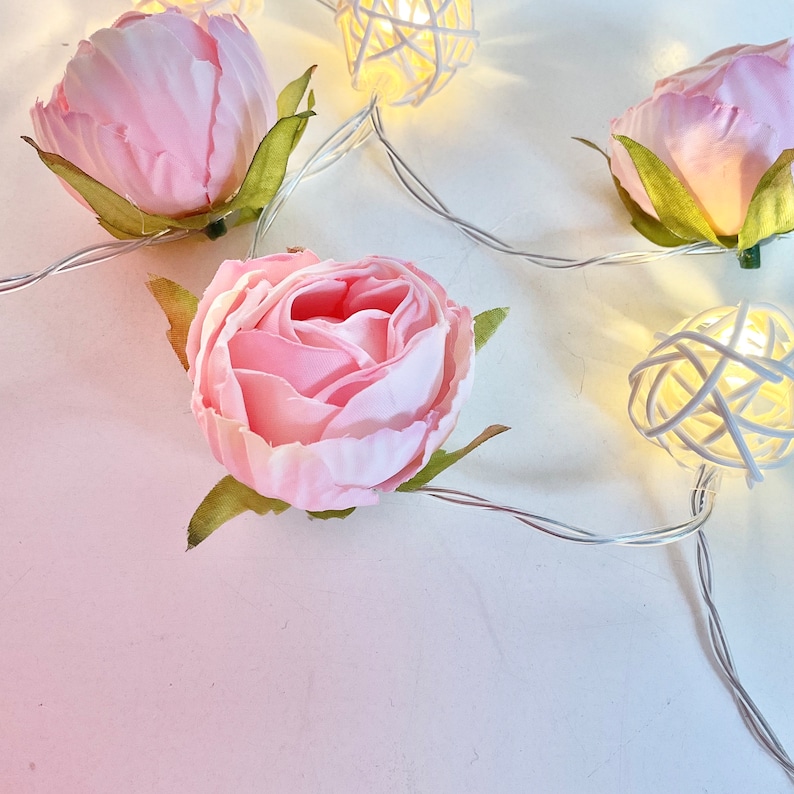 May include: Pink fabric rose flower buds with white woven ball lights on a white background. The lights are strung together with a silver wire.