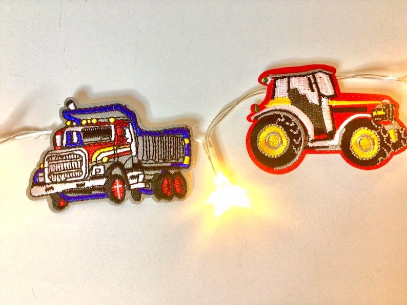 May include: A string of fairy lights with a red and yellow tractor and a blue and red dumper truck attached. The lights are white and the truck and tractor are embroidered.