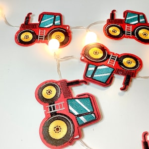 May include: A string of red toy tractor lights with white lights. The tractors have black wheels, yellow rims, and a teal cab.