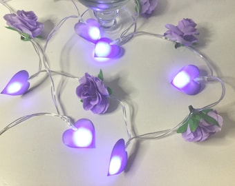 May include: Purple fairy lights with heart-shaped bulbs and purple fabric flowers. The lights are strung together with clear wire.