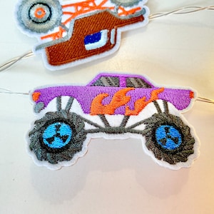 May include: Embroidered monster truck patch with a purple body, orange flames, and black wheels. The wheels have a blue center. The patch is white with a gray border. A second patch of a monster truck is visible in the background.