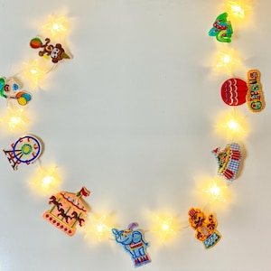 Puede incluir: Una cadena de luces blancas con decoraciones coloridas de temática circense, que incluyen una noria, un carrusel, un elefante, un payaso y un cubo de palomitas. Las luces están conectadas con un cable transparente.