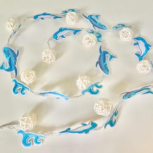 May include: A string of decorative lights featuring blue and white dolphin and wave designs. The string lights also include white woven sphere lights. The lights are strung on a silver wire.
