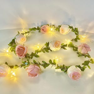 May include: A floral garland with pink and white roses, green foliage, and small star-shaped lights. The fairy lights are arranged in a circular pattern, providing a warm, inviting glow. Suitable for weddings, parties, or home decoration.