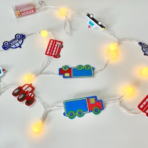 May include: String lights with warm white LEDs and vehicle-shaped decorations. The decorations include a red bus, blue police car, and red truck. The lights are powered by a battery pack.