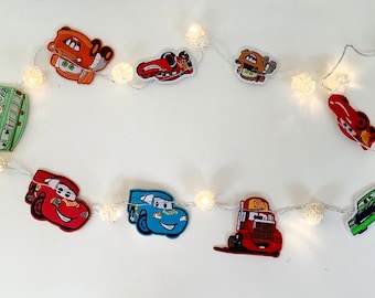 May include: A string of white fairy lights with white cotton ball-shaped bulbs. Each bulb has a different Cars character attached, including Lightning McQueen, Mater, and Tow Mater. The lights are battery-operated and have a Kodak battery pack.