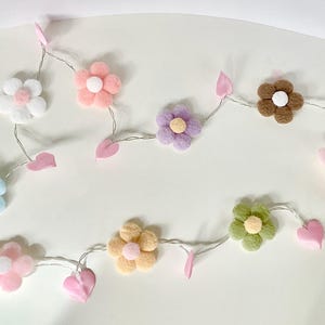 May include: A string of decorative lights featuring colorful felt flowers in shades of pink, blue, yellow, green, brown, and purple. Each flower is connected by a thin wire and includes pink felt leaves and hearts.