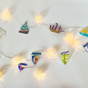 Peut inclure: Une guirlande de lumières blanches avec des découpes en papier de voiliers de différentes couleurs et motifs. Les lumières sont accrochées à un mur blanc.