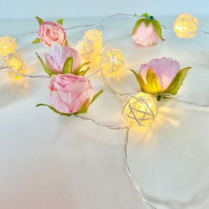 May include: A string of white fairy lights with pink fabric flowers and white woven balls. The lights are strung together with a clear wire.