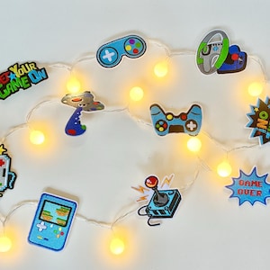 May include: A string of white fairy lights with colorful game-themed patches. The patches include a joystick, a game controller, a spaceship, and the text "Get Your Game On!" and "Game Over".