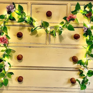 May include: A cream-colored dresser with brown knobs is adorned with a garland of faux green leaves, pink, purple, and white flowers, and tiny fairy lights. The garland is draped across the top of the dresser.