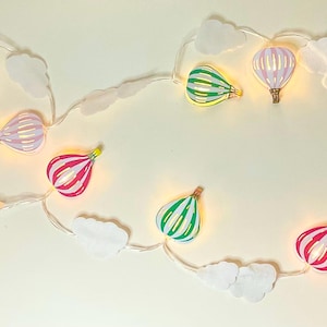 Op de afbeelding: Een string met lichtsnoeren met lampjes in de vorm van luchtballonnen. De lampjes zijn in verschillende kleuren, waaronder geel, blauw, roze, groen en rood. De lampjes zijn aan een wit snoer geregen met witte wolkvormen tussen de lampjes. De lampjes werken op batterijen.