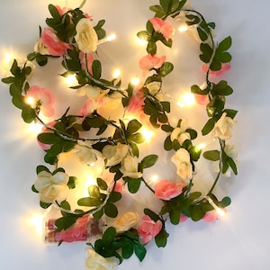 Puede incluir: Una guirnalda de luces de hadas adornada con rosas artificiales rosas y crema y hojas verdes. Las luces están encendidas, iluminando las flores. Un paquete de baterías es visible en la parte inferior.