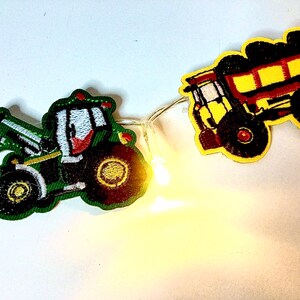 May include: A string of lights with two felt patches, one of a green tractor and one of a yellow dumper truck. The tractor has a red cab and a white windscreen. The dumper truck has a red bed and black tyres.