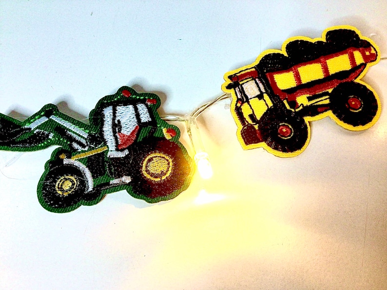 May include: A string of lights with two felt patches, one of a green tractor and one of a yellow dumper truck. The tractor has a red cab and a white windscreen. The dumper truck has a red bed and black tyres.