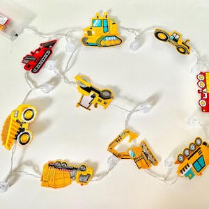 May include: A string of fairy lights featuring colorful construction vehicle designs. The lights include yellow, red, and blue trucks, excavators, and other equipment. The lights are strung on a clear wire with small, clear bulb accents.