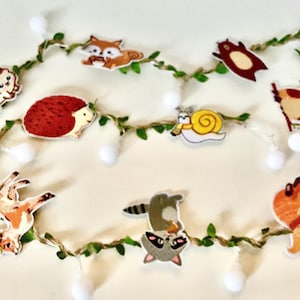 May include: A whimsical string of lights featuring woodland animal cutouts, including a hedgehog, fox, and raccoon. The garland is adorned with green leaves and small white pom-poms, creating a charming, nature-inspired decoration.