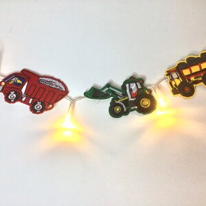 May include: A string of fairy lights with red, green, and yellow construction vehicle cutouts. The vehicles include a dumper truck, a tractor, and a pick-up truck.