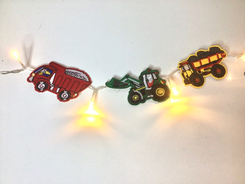 May include: A string of fairy lights with red, green, and yellow construction vehicle cutouts. The vehicles include a dumper truck, a tractor, and a pick-up truck.