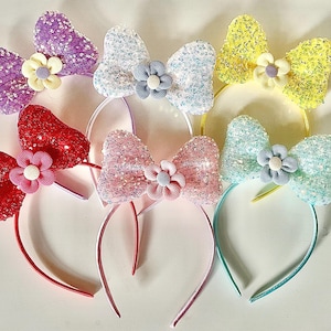 Puede incluir: Una colección de diademas coloridas con lazos de lentejuelas y flores de tela. Las diademas presentan lazos en morado, blanco, amarillo, rojo, rosa y aguamarina, cada uno con una flor a juego.