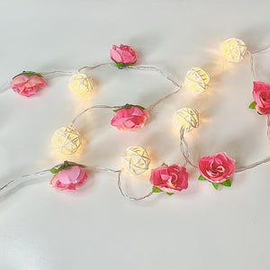 May include: A string of white lights with pink fabric flowers. The lights are in the shape of a ball and are strung together with a clear wire.