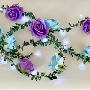 May include: A decorative string of lights featuring artificial purple and light blue roses intertwined with green leafy vines. The garland includes small, bright white LED lights spaced along the wire, creating a floral illumination effect.