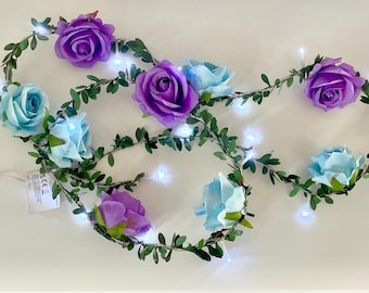 May include: A decorative string of lights featuring artificial purple and light blue roses intertwined with green leafy vines. The garland includes small, bright white LED lights spaced along the wire, creating a floral illumination effect.