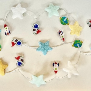 Puede incluir: Una cadena de luces decorativa con adornos temáticos del espacio. La cadena incluye luces suaves en forma de estrella de color azul claro, amarillo y blanco, junto con pequeñas naves espaciales, planetas y adornos con forma de astronauta.