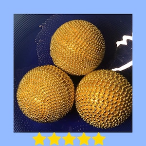 Puede incluir: Tres adornos redondos de color dorado con una superficie texturizada. Los adornos están dispuestos en forma de triángulo sobre un fondo azul. 5 de 5 estrellas.