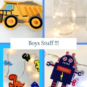 May include: A set of two glass jars with fairy lights inside. The jars are decorated with pom poms and fabric appliques. One jar has a blue dinosaur applique and the other has a yellow construction truck applique. The text "Boys Stuff!!!" is written below the jars.