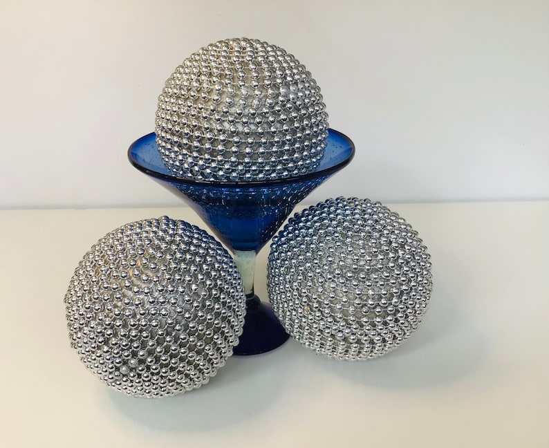 K&ouml;nnte beinhalten: Drei silberne Dekorationssph&auml;ren, jede mit kleinen, runden Perlen bedeckt, werden auf einem blauen Glaspodest pr&auml;sentiert. Die Sph&auml;ren sind in einer dreieckigen Formation angeordnet, wobei die oberste Kugel auf dem Podest ruht und die beiden unteren Kugeln auf dem Tisch liegen.