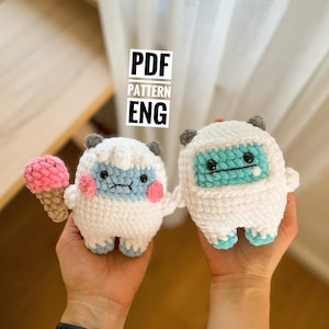 May include: Two crocheted amigurumi toys, one white with blue and pink details holding a pink and brown ice cream cone, and the other white with blue details. The toys are being held by a person's hands. The text "PDF PATTERN ENG" is visible in the image.
