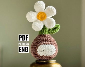 May include: Crocheted amigurumi flower pot with a white flower and a brown face. The pot is sitting on a gold metal stand.