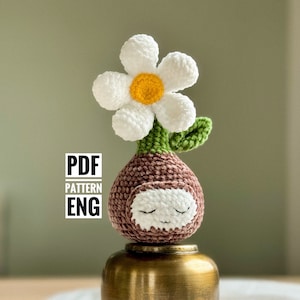 Pode incluir: Pote de flores de amigurumi crochetado com uma flor branca e um rosto marrom. O pote está em um suporte de metal dourado.