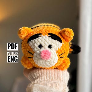 May include: Crocheted stuffed toy of a tiger with orange and black stripes, white face, and pink nose. The toy is being held in a hand. The text "PDF PSTTERN ENG" is visible in the image.