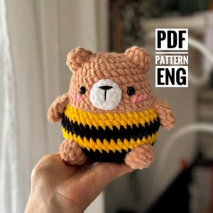 Può includere: Un orsetto all'uncinetto fatto a mano con un design ispirato alle api. L'orsetto presenta una testa e arti color marrone chiaro, un corpo a strisce nere e gialle e un muso bianco con un naso nero. Il testo "PDF PATTERN ENG" è visibile.