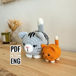 May include: Two crocheted cat plushies, one grey with black stripes and white accents, the other orange with black stripes and white accents. The plushies are sitting on a wooden surface. The text "PDF PATTERN ENG" is visible in the image.