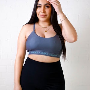 Può includere: Una donna indossa un reggiseno sportivo grigio con la scritta "AURORA" stampata sulla fascia, abbinato a leggings neri e grigi. Il reggiseno ha spalline sottili e uno scollo rotondo. I leggings hanno un design a blocchi di colore. Lo sfondo è un muro di mattoni bianchi.